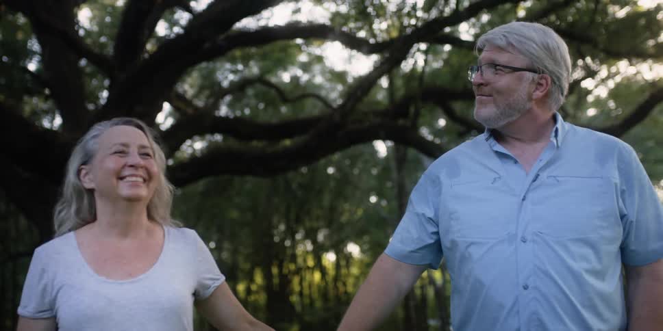 An elderly man and woman talk and hold hands as they walk by large trees