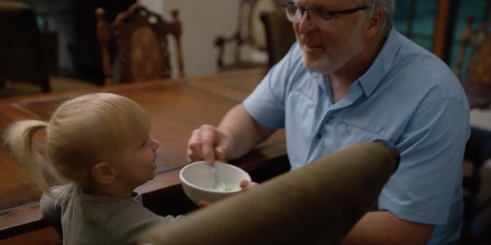 A man feeding a little girl with a spoon and bowl next to a table