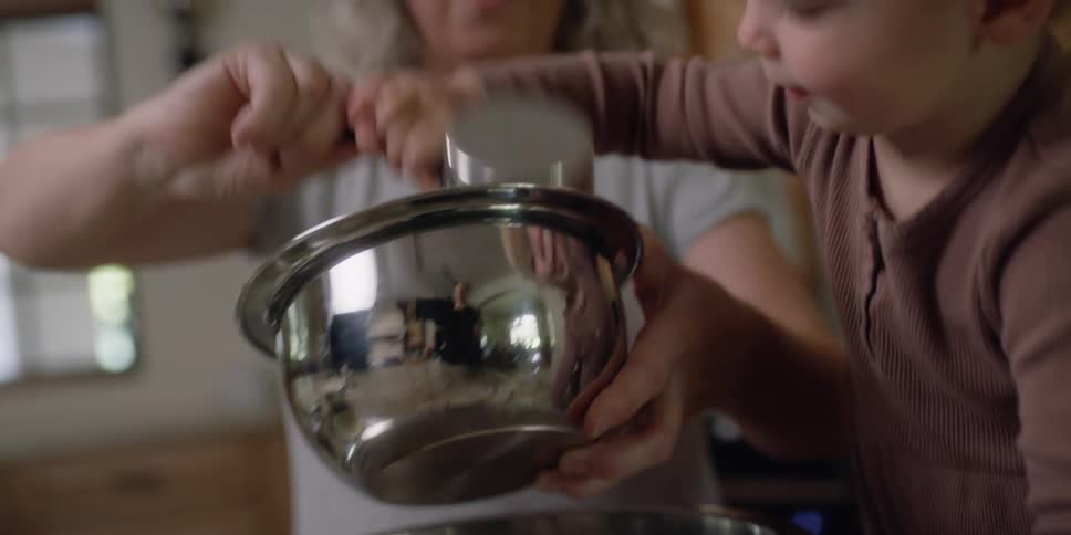 A toddler helps her grandmother add ingredients in a mixing bowl