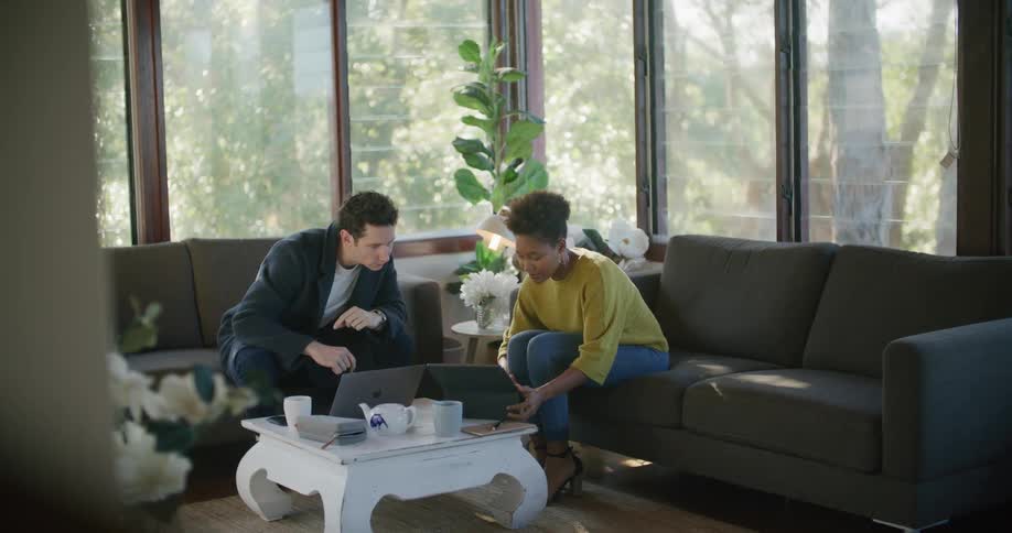 A man leans forward to look at the screen of the laptop of a woman sitting on a sofa opposite him in the living room of a house