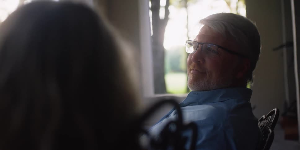 A man and a woman in a room sitting on chairs with a wall and window in the background