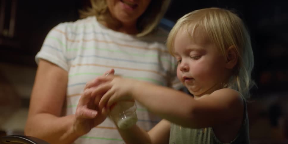 A young toddler shakes seasoning from a container with a woman standing close beside