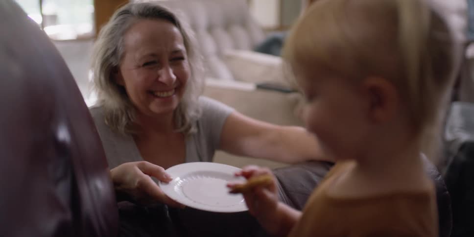 A woman kneels beside a toddler girl eating food