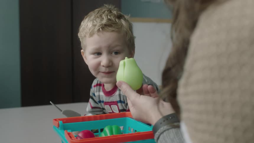 Woman holds up a toy pear to a young toddler who is holding toy knives