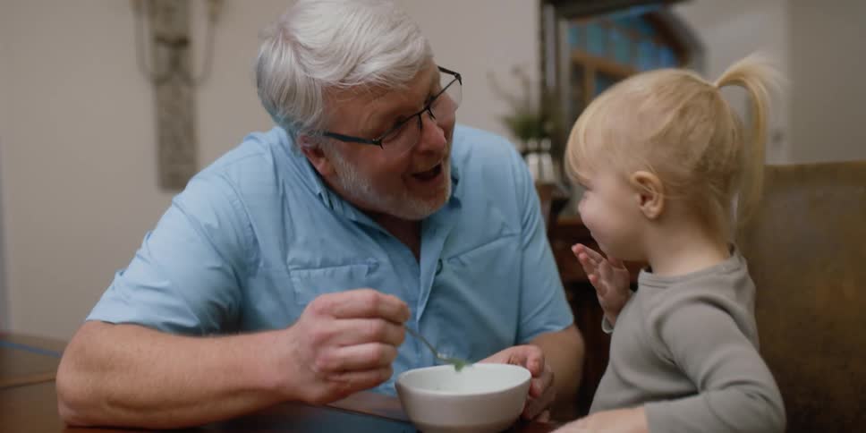 A grandfather smiles at granddaughter after feeding her ice cream at kitchen table