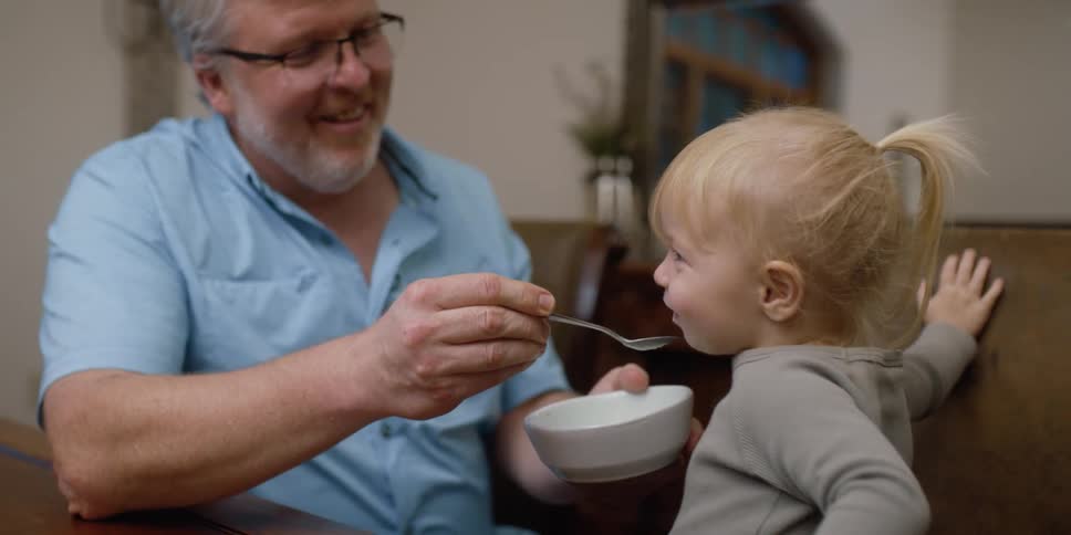 A man feeding a little girl with a bowl and spoon next to a table