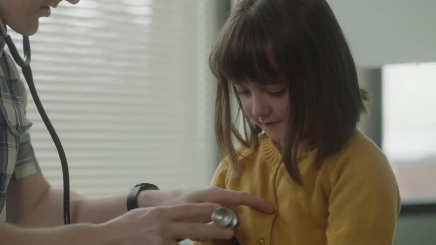 A doctor puts a stethoscope to his ear and to the chest of a young girl patient