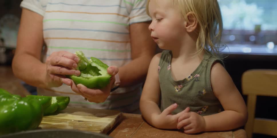 Grandmother takes the seeds out of green bell peppers next to little granddaughter in the kitchen
