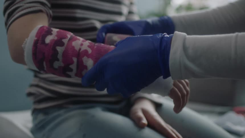 A person with gloves in a clinic wrapping a girls hand with a bandage