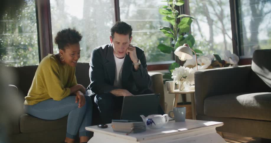 Two coworkers are sitting on a couch talking and smiling and pointing at laptop