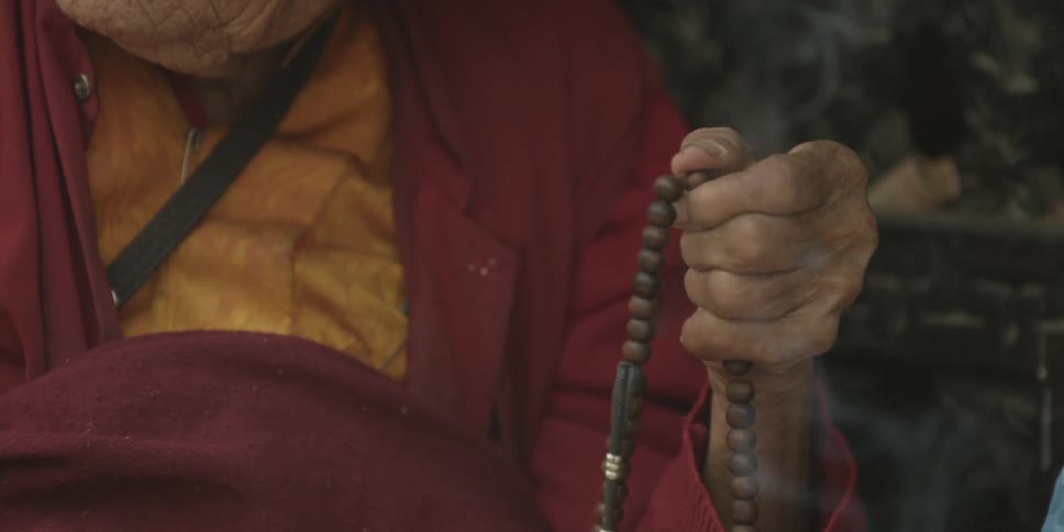 An elderly woman clutches a string of beads outside in front of smoke