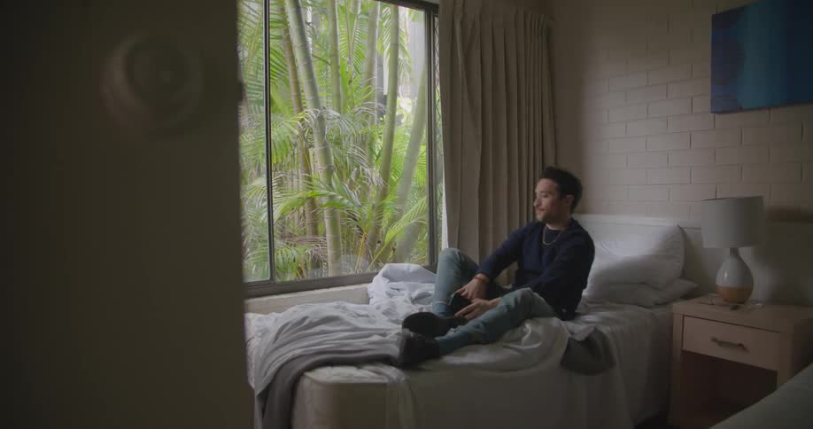 Man sits on a messy bed and looks at his cellphone then he lays back on the bed
