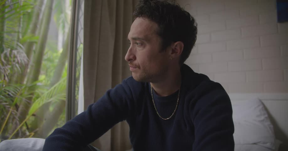 Man sits on bed frowning and looking upset before scrolling on mobile phone