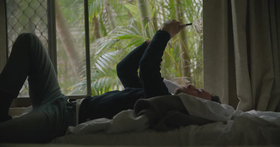 Man lies on his back on bed looking at mobile phone