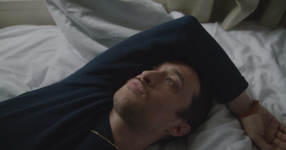 Man lies down onto bed sighing as he puts hand over forehead on white bedspread