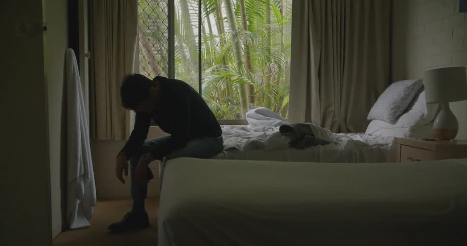 A man in a bedroom sitting on a bed and looking at a smart phone with a wall and window in the background