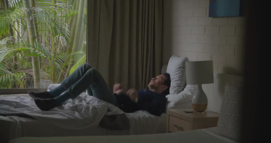 A man sitting in bed looking at a smart phone with a wall and window in the background
