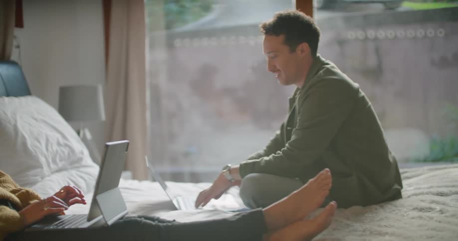 A man and woman sitting in bed each working on laptops and talking