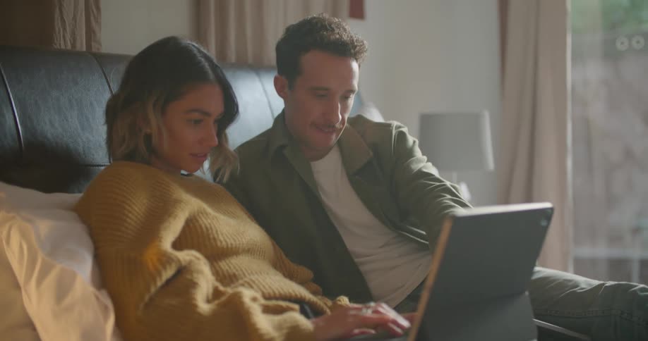 Couple sit in bed talking as woman types on laptop