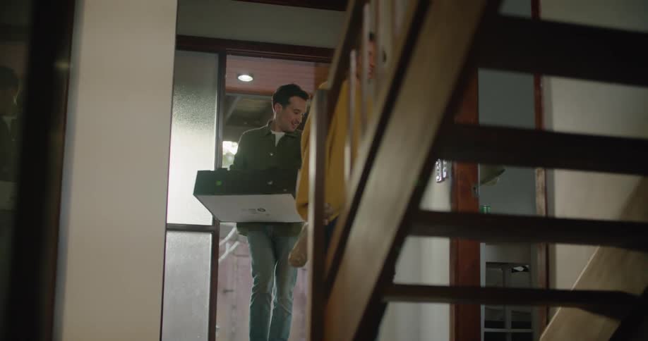 A woman opens the door and she and her husband walk up the staircase carrying a box and a bottle in a paper bag