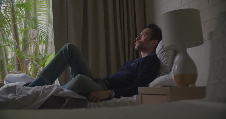 A man sitting in bed looking at a smart phone with a wall and window in the background