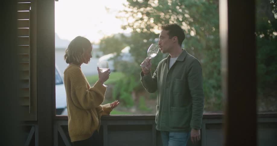 A man and a woman on a porch drinking from glasses with trees the sky and clouds in the background