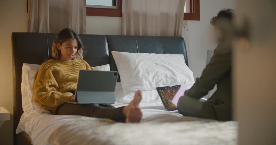 A man and a woman in a room sitting on a bed looking at laptops