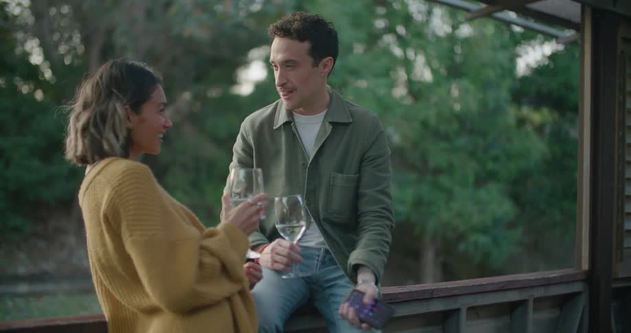 A man with a smart phone and a glass sitting on the porch and talking to a woman with trees the sky and clouds in the background