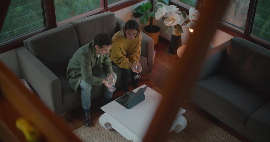 Man and woman sit on a sofa talking while holding wine glasses and looking at a laptop