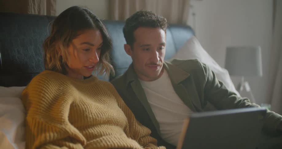 Man and woman recline in bed with padded headboard talking as they view laptop
