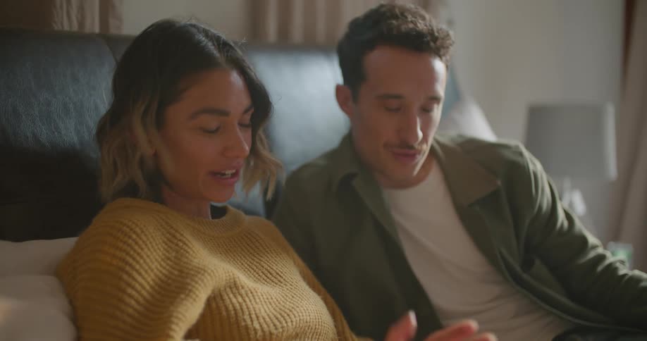 Man and woman lay on a bed and smile and laugh while looking at a laptop screen