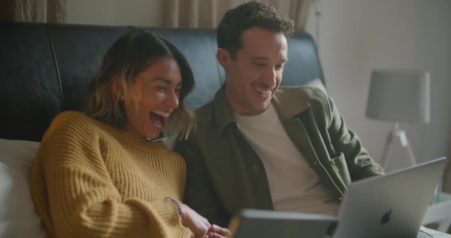 A man and a woman in a room sitting on a bed looking at laptops