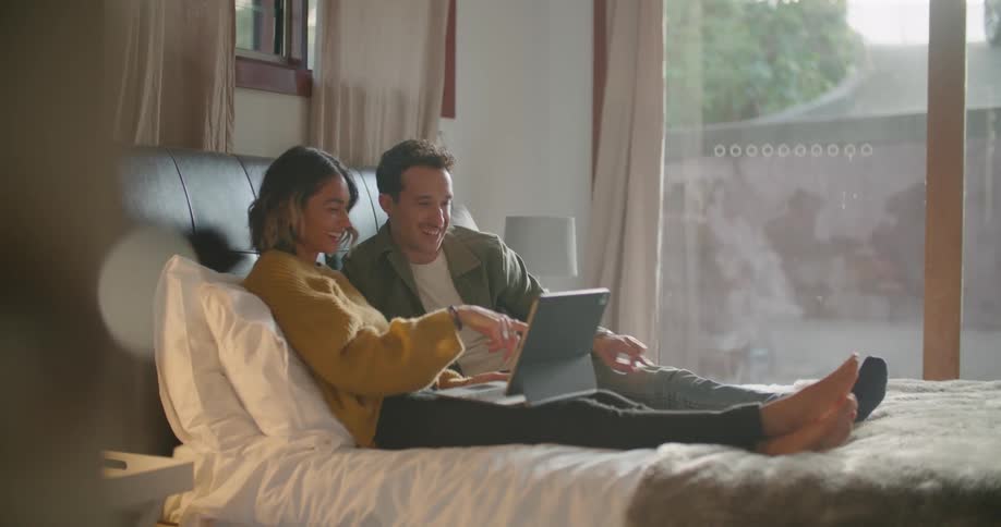 Couple reclines on bed laughing and talking as they view tablet