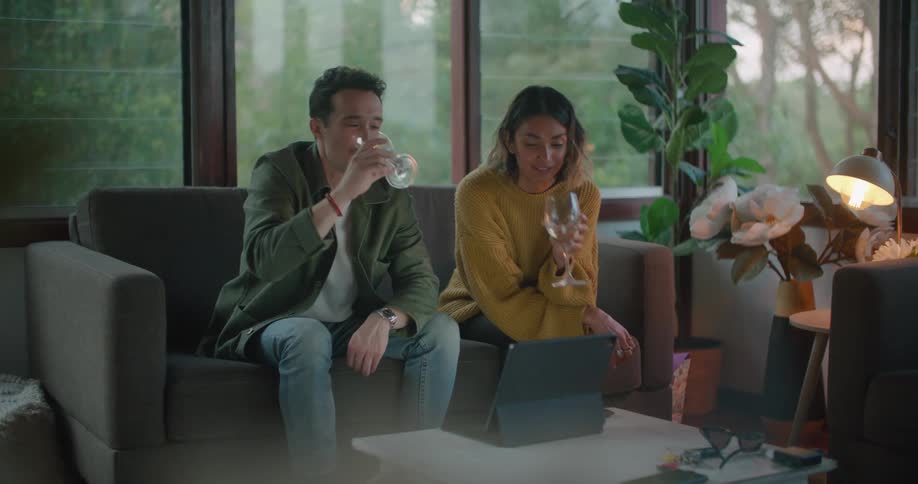 A man and woman sitting on a love seat clink their glasses and drink wine