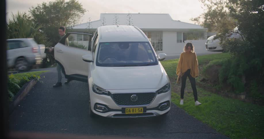 A man and a woman coming out of vehicle in a driveway with a house trees the sky and clouds in the background