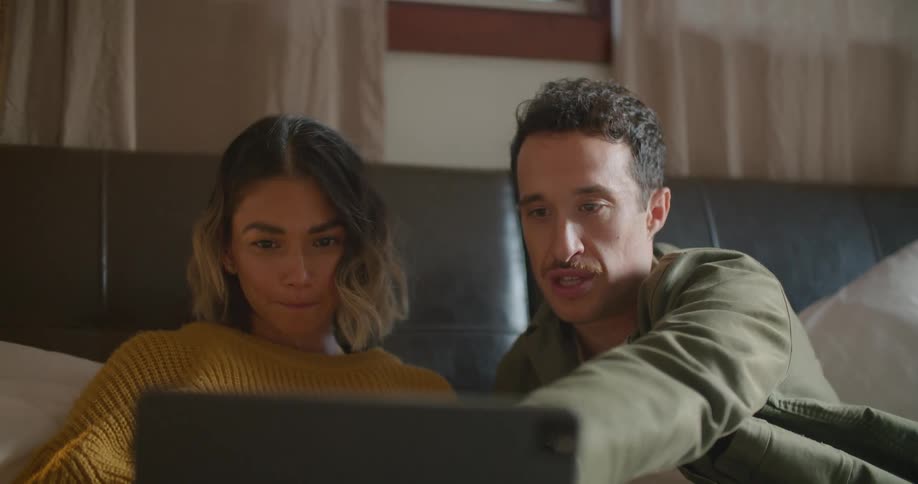 A man points at the screen of a laptop on the lap of a woman sitting beside him indoors during the day