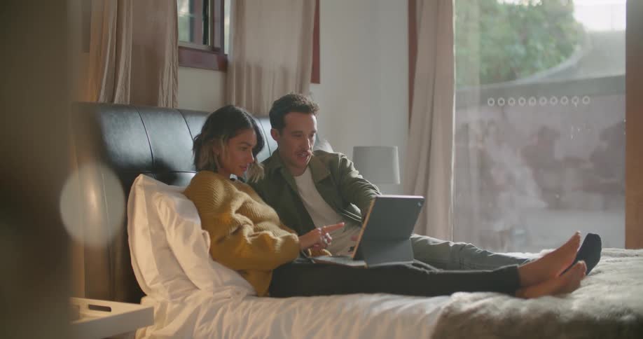 A man and a woman in a room laying on a bed looking at a laptop