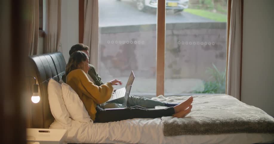 A man and a woman in a room sitting on a bed looking at laptops