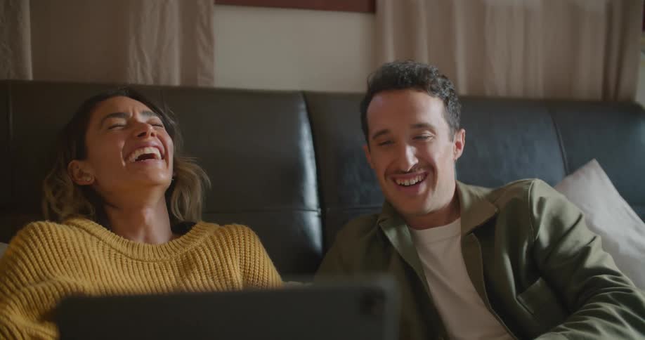 Man and woman sit on a sofa and laugh as they look at a laptop screen