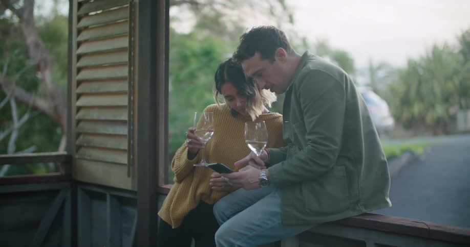 Man in green jacket and jeans and woman wearing yellow sweater hold wine glasses and use smartphone