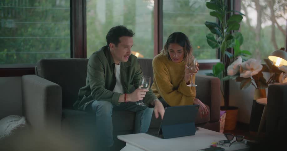 A man and woman sit side by side on a love seat holding wine glasses and looking down at a laptop as they talk