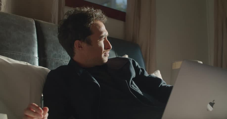 A man twirls a pen while lost in thought as he sits on a bed