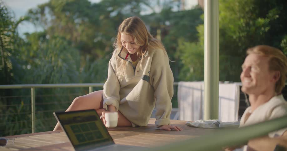 A woman sits on a table outdoors drinking coffee while her partner works on his laptop