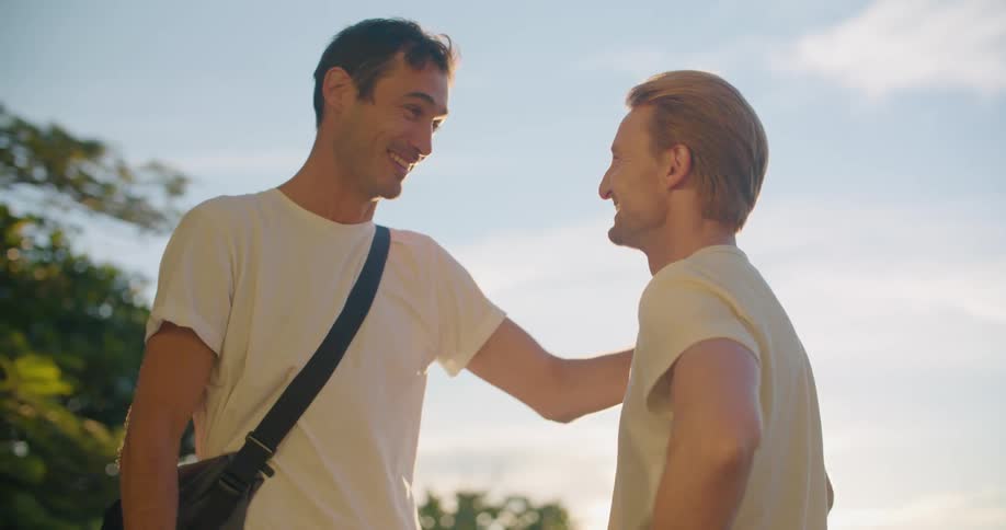 Two men stand outside laughing and talking as one touches the others shoulder