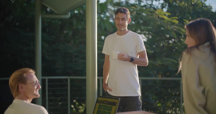 A man sits with his laptop outside while chatting with another man and a woman on their balcony