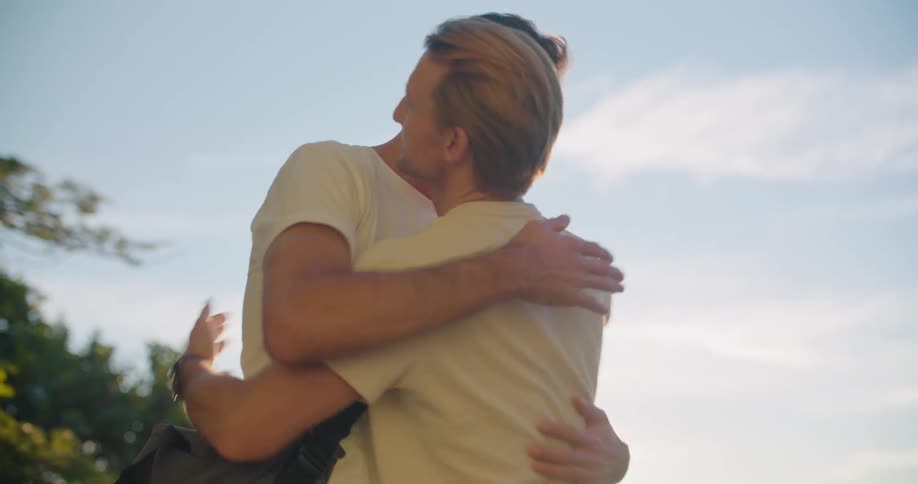 Men hug in greeting then stand talking outdoors on sunny day
