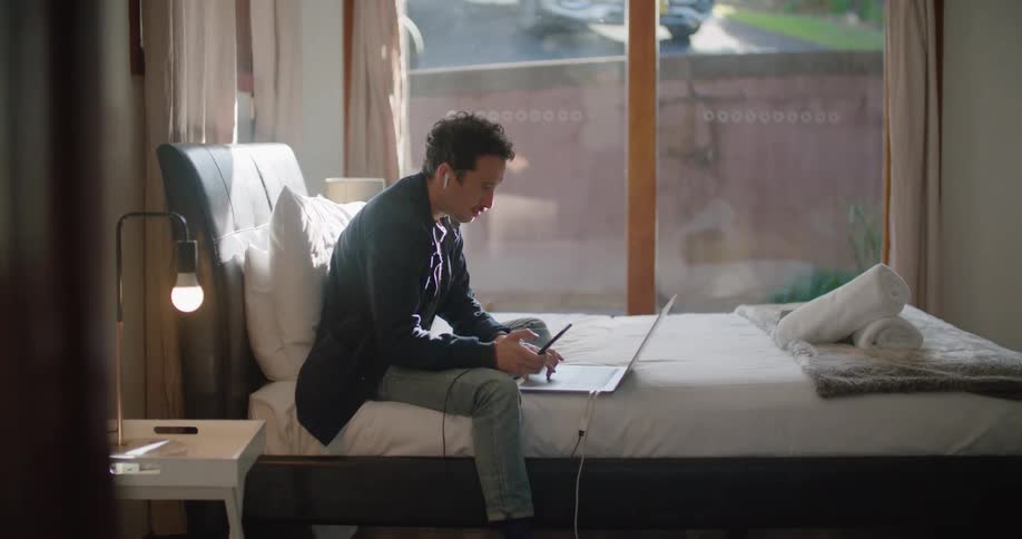 Man sits on a bed and scrolls on his laptop
