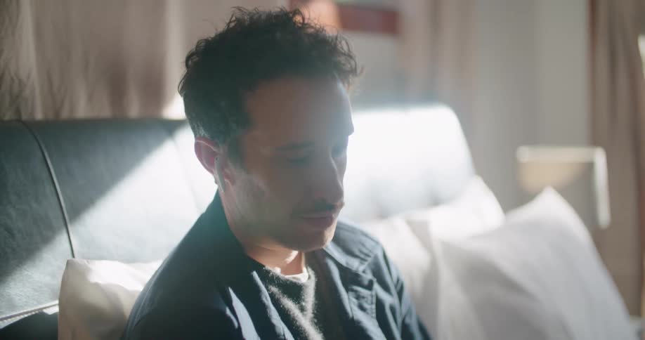 Man sits on a bed with a laptop and holds a cellphone as he talks
