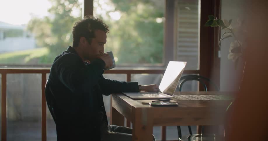 A man drinks coffee while using his laptop and working from home at his kitchen table