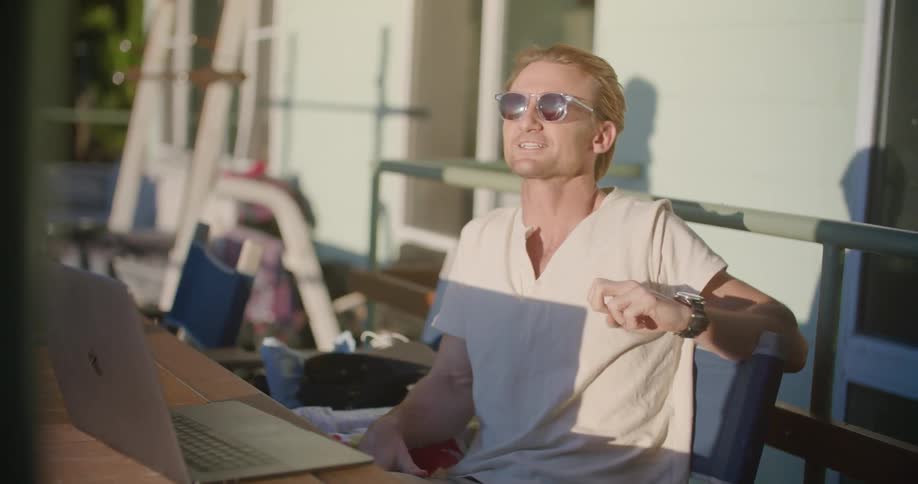 A man sitting outdoors in front of a laptop on a wooden table talks to someone out of frame